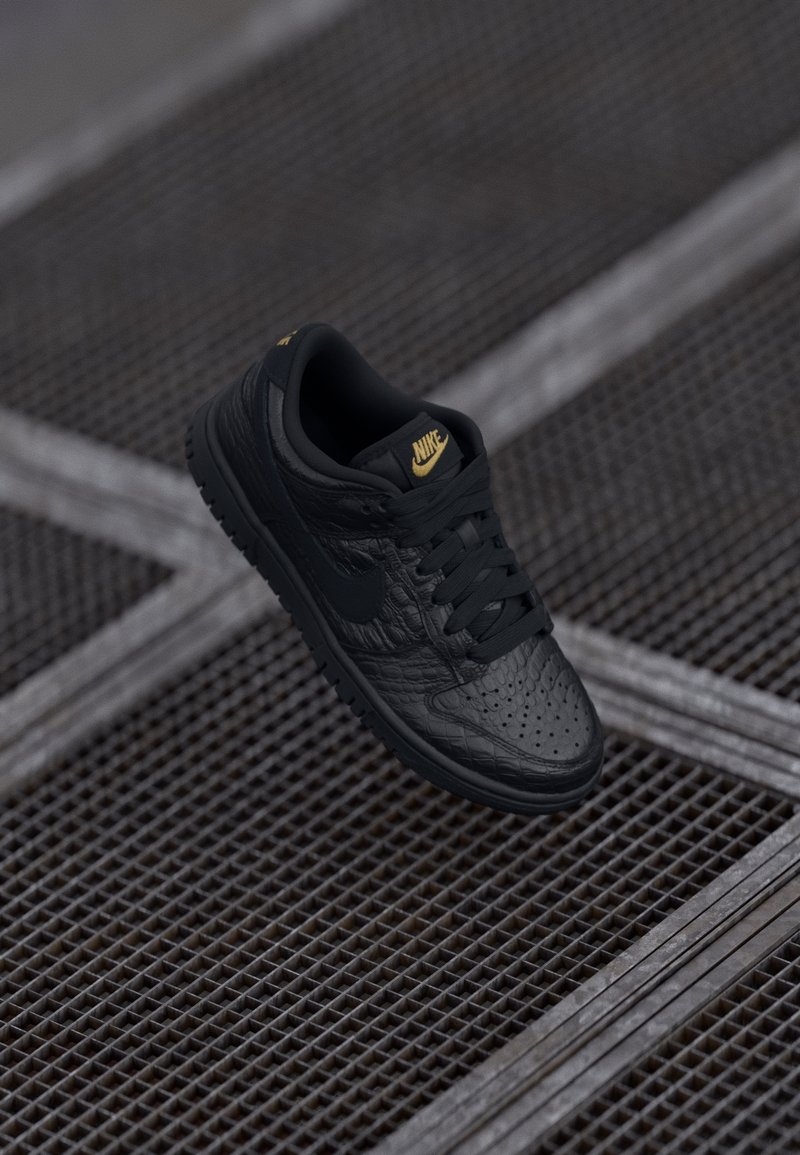 Baskets Nike en cuir noir texturé avec logo doré sur la languette, flottant au-dessus d'une surface en grille métallique.