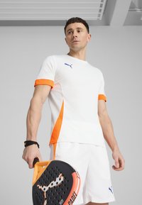 Camiseta deportiva blanca con detalles en naranja y un logo azul, acompañada de pantalones cortos blancos. Sosteniendo una pala de pádel negra y naranja.