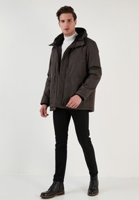 Brun isoleret jakke med hætte, der har lynlås foran, sort fleeceforing og elastiske manchetter, parret med sorte slim-fit bukser og støvler.