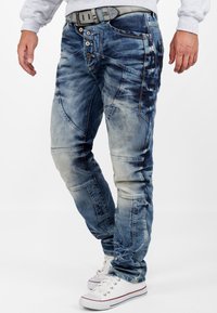 Jeans en denim bleu avec un design décoloré, plusieurs boutons à l'avant, des coutures accentuées et une coupe slim, associés à des baskets en toile blanches.