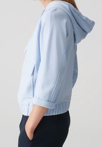 Žena v světle modré mikině s kapucí, s vyhrnutými rukávy a přední kapsou, v tmavých kalhotách, stojící v profilu.