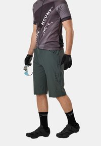 Homme portant un maillot de cyclisme gris à manches courtes, un short de cyclisme vert foncé, des gants noirs, des chaussettes et des chaussures, tenant des lunettes de soleil réfléchissantes dans la main gauche.