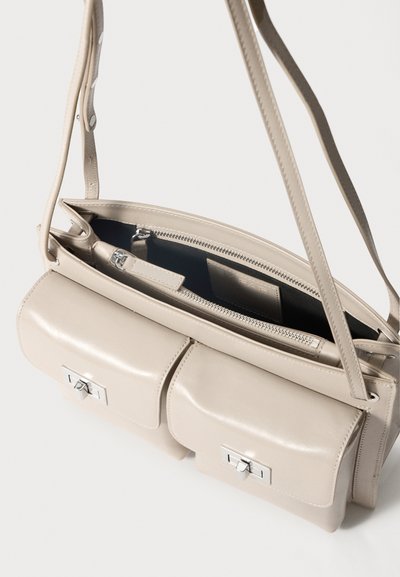 Beige nahk käekott, millel on kaks esikotit, hõbedased detailid ja lukuga peamine osa; sile tekstuur ja reguleeritav õlavarre rihm.