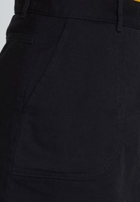 Pantaloni di cotone neri con una texture liscia, cuciture diagonali e una tasca laterale. La vita è piatta e rinforzata.