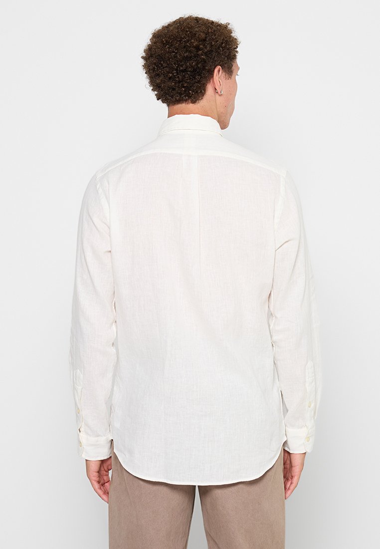 Chemise à manches longues beige clair en lin. Elle présente un col classique et des poignets boutonnés, avec une coupe droite et un détail de couture au dos.