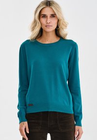 Kobieta o blond włosach do ramion, ubrana w tealowy sweter z długim rękawem i ciemne brązowe spodnie, stojąca na tle jednolitym.