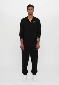 Sort zip-up trøje med lange ærmer, der har et lille logo, parret med matchende sorte joggers og hvide sneakers. Enkel, strømlinet design.