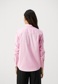 Pitkähiha, nappikauluspaita kirkkaan pinkistä puuvillasta, jossa on klassinen kaulus, suora helma ja napitetut hihansuut. Yhdistetty laskostettuihin mustiin housuihin.