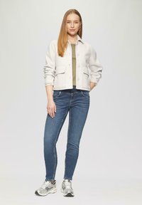 Veste boutonée gris clair avec deux poches avant, portée sur un haut vert, associée à un jean skinny bleu foncé et des baskets argentées.