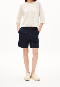 Marinblå shorts med knäppning och fickor, tillsammans med en lös, krämfärgad stickad topp och ljusa slip-on-skor.