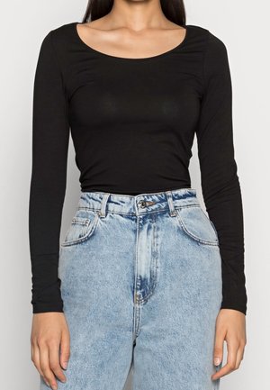 Woman wearing a fitted black long-sleeve top tucked into high-waisted light blue denim jeans against a white background.