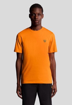 Orange bomull t-shirt med korta ärmar, rund halsringning och en liten svart logotyp på vänster bröst. Bärs med svarta byxor.