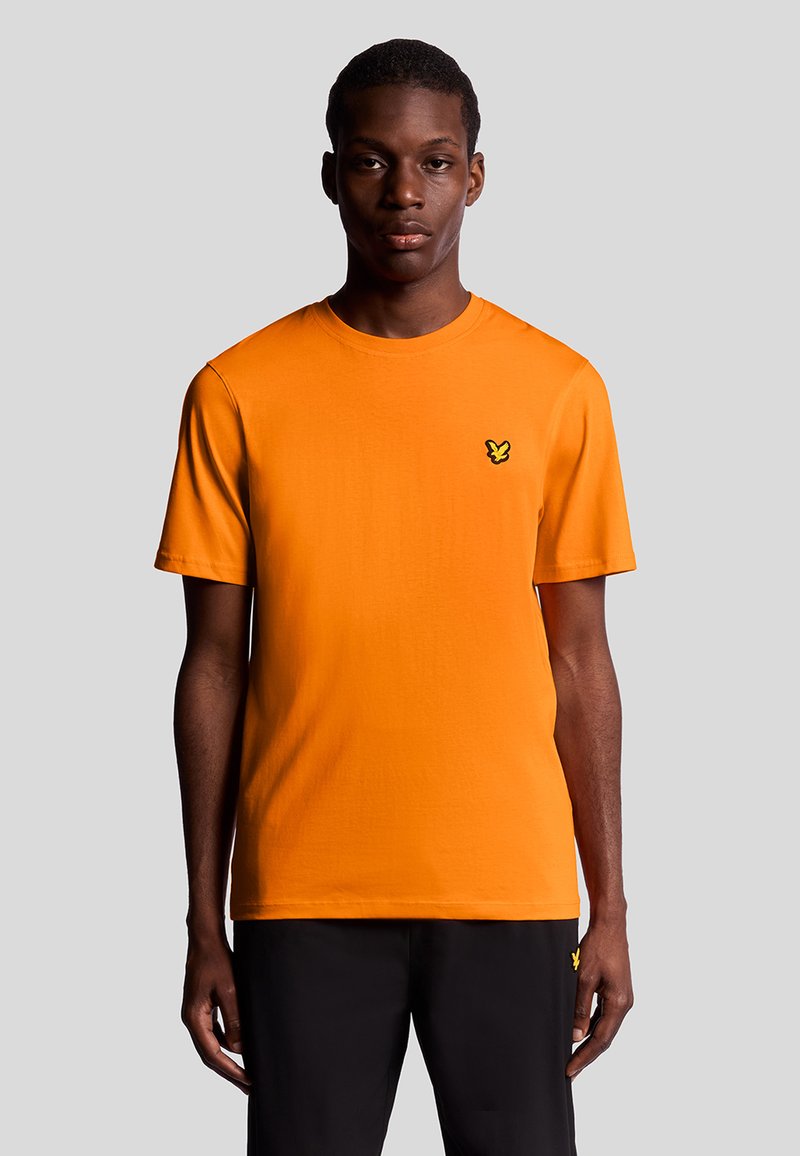 T-shirt di cotone arancione con maniche corte, scollo rotondo e un piccolo logo nero sul lato sinistro del petto. Indossata con pantaloni neri.