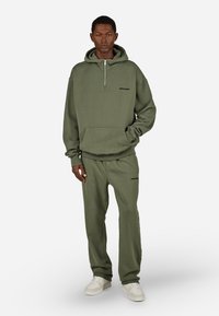 Χακί φούτερ με μισό φερμουάρ και τσέπη καγκουρό, συνδυασμένο με παντελόνι σε αντίστοιχο χρώμα. Το ύφασμα είναι μαλακό, από μείγμα βαμβακιού, με ελάχιστο branding.