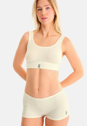 Jeune femme portant un soutien-gorge de sport en tissu côtelé crème et un short assorti avec de petites étiquettes logo sur fond blanc uni.