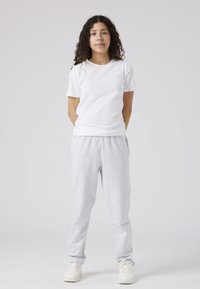 Witte t-shirt, lichtgrijze sweatpants en witte sneakers. Model staat rechtop tegen een effen achtergrond, met de armen ontspannen langs het lichaam.