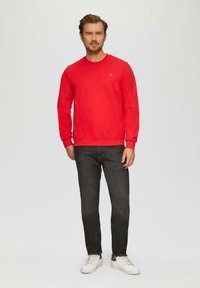 Sweatshirt rouge avec un col rond, associé à un jean foncé et des baskets blanches. Le sweatshirt présente un petit logo sur la poitrine.