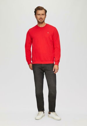 Rode sweatshirt met een ronde hals, gecombineerd met donkere jeans en witte sneakers. De sweatshirt heeft een klein logo op de borst.