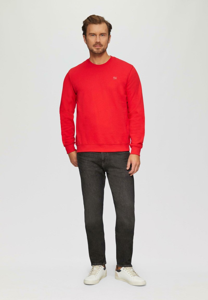Sweatshirt rouge avec un col rond, associé à un jean foncé et des baskets blanches. Le sweatshirt présente un petit logo sur la poitrine.