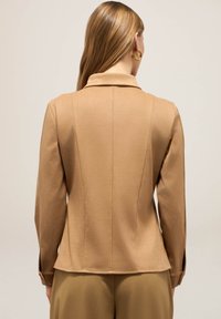 Veste beige avec un design structuré, col cranté et manches longues. Tissu lisse avec des coutures décoratives et des fentes sur les côtés en bas.