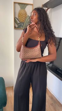 Mulher com cabelos longos e encaracolados, vestindo um traje preto, segura uma bolsa de padrão castanho no interior, perto de uma obra de arte de ananás.