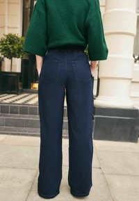 Pantaloni de denim largi de culoare albastră închisă, cu talie înaltă, având două buzunare pe spate și un pulover texturat verde deasupra.