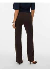 Temno rjave hlače z ravnimi hlačnicami iz elastičnega blaga. Imajo gladko teksturo in oprijeto pasnico, nošene z rjavimi čevlji z višjo peto.