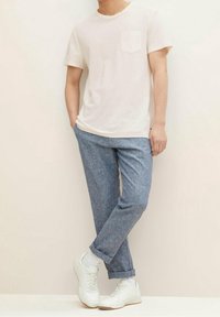 Osoba w białej koszulce z krótkim rękawem i kieszonką, niebieskich spódnicach podwiniętych u dołu, białych skarpetkach i białych sneakersach, stojąca przy gładkiej ścianie.