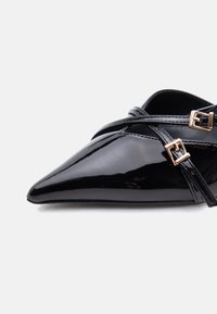 Γυαλιστερό μαύρο γυναικείο παπούτσι με μυτερή μύτη και επίπεδη σόλα, με δύο λεπτές λουρίδες που διασταυρώνονται και κλείνουν με μικρές χρυσές τετράγωνες αγκράφες.