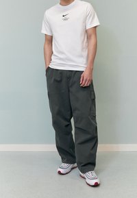 Vit bomullströja med svart Nike-logotyp och text; grå cargobyxor med sidofickor; vita sneakers med röda accenter.