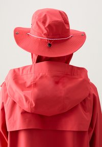 Manteau imperméable rouge avec une grande capuche flexible et un chapeau seau assorti. Il dispose d'un cordon de serrage et d'accents de boutons pour un ajustement et un style personnalisables.