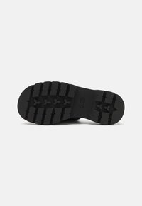 Semelle en caoutchouc noire d'une chaussure avec une bande de roulement profonde et des crampons rectangulaires et triangulaires pour l'adhérence sur un fond clair uni.