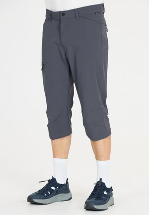 Tummanharmaat polvipituisiin shortseihin sivutaskussa vetoketju, valkoiset sukat ja tummansinivihreät vapaa-ajan kengät vaalealla taustalla.