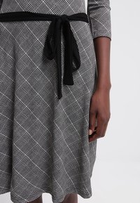 Rutig klänning i svart och vitt, 3/4-lång ärm, åtsittande överdel, utsvängd kjol. Har ett svart bälte i midjan och en mjuk tygtextur.