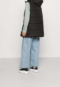 Svart vadderad väst med huva, kombinerad med en ljusgrön långärmad tröja, vida ljusblå jeans och svarta sneakers med vita sulor.