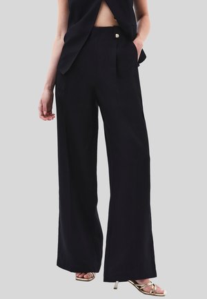 Schwarze weit geschnittene Hose aus glattem Stoff, mit einer vorderen Falte und einem dekorativen Knopfdetail, kombiniert mit einem eleganten Oberteil und Sandalen.