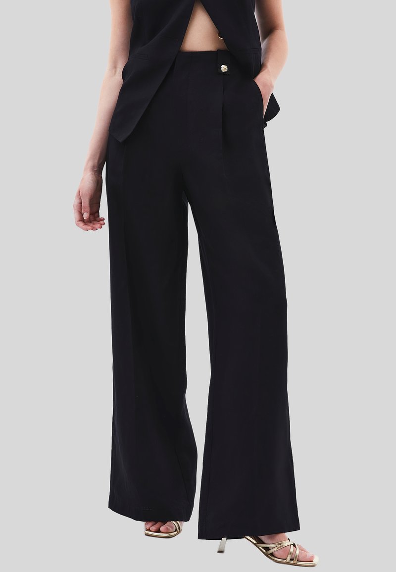 Zwarte wijde broek gemaakt van soepel materiaal, met een voorplooien en een decoratieve knoopdetail, gestyled met een elegante top en sandalen.