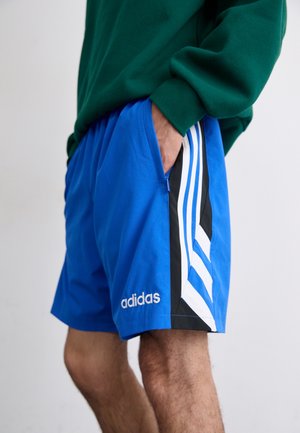 Personne portant un short Adidas bleu avec des bandes blanches et des panneaux noirs, main dans la poche, assorti à un sweat-shirt vert.