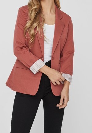 Vrouw draagt een roestkleurige blazer met opgerolde gestreepte manchetten, een wit topje en zwarte broek, en past haar linkermouw aan met haar rechterhand.