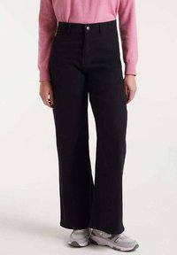 Persona vestida con pantalones anchos de cintura alta negros, blusa de manga larga rosa y zapatillas gris y blancas, de pie contra un fondo liso.