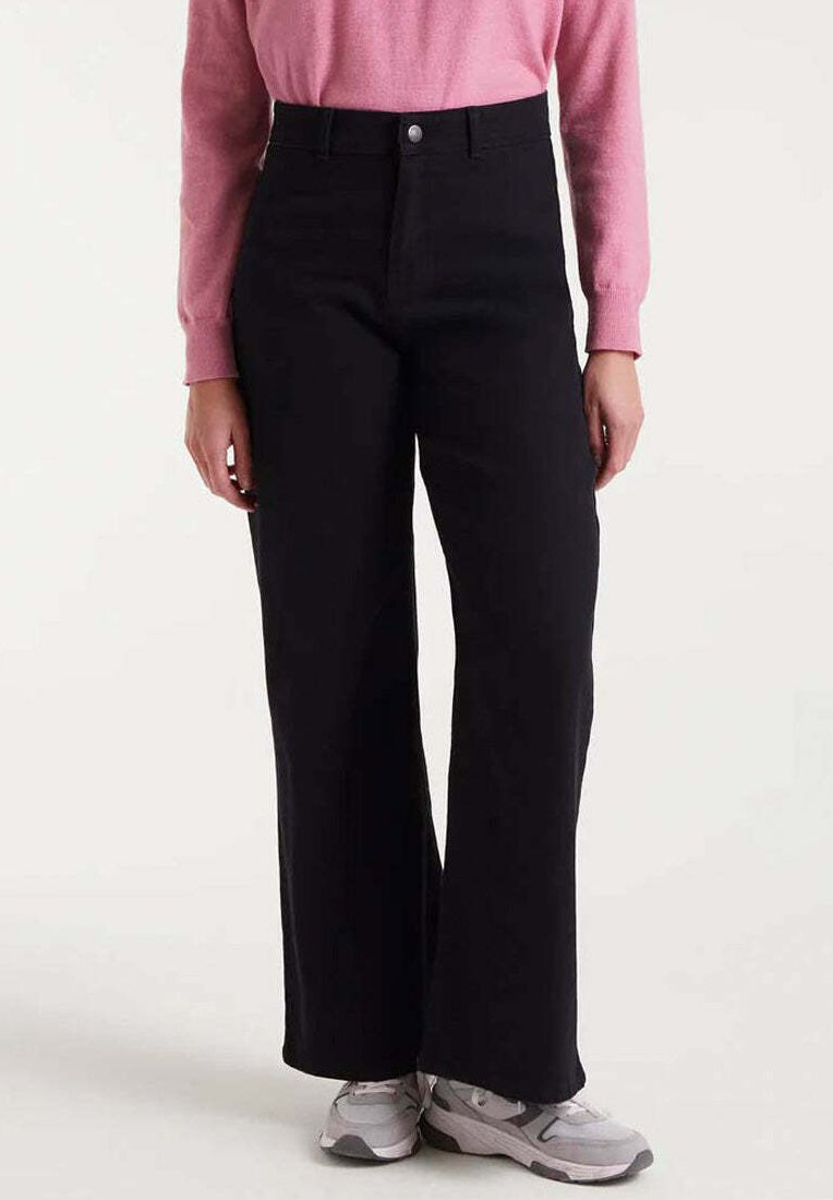 Persona vestida con pantalones anchos de cintura alta negros, blusa de manga larga rosa y zapatillas gris y blancas, de pie contra un fondo liso.