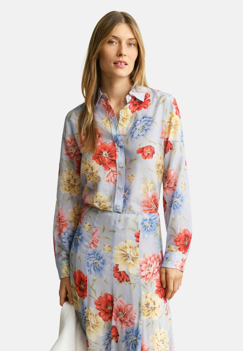 Femme aux longs cheveux blonds portant une chemise boutonnée et une jupe assorties bleu clair à motifs floraux rouges, jaunes et bleus, tenant un objet blanc.