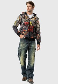 Kapuzensweatshirt mit Reißverschluss und grafischem Design in Schwarz, Rot und Gelb; kombiniert mit blauen Cargo-Jeans, die über mehrere Taschen und distressed Akzente verfügen.