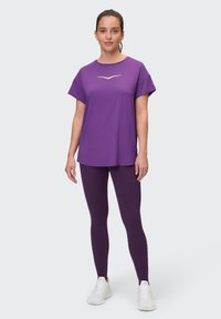 Lila Sportshirt mit kurzen Ärmeln und einem goldenen Logo, kombiniert mit passenden lila Leggings und weißen Schuhen, auf einem neutralen Hintergrund.