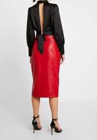 Sort satinbluse med udskæring på ryggen, bundet i taljen, kombineret med en tætsiddende rød lædermidi-nederdel og sorte højhælede sandaler.