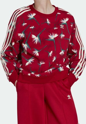 Person trägt roten Adidas-Sweatshirt mit weißem Blumenmuster und drei weißen Streifen an den Ärmeln, kombiniert mit passenden roten Adidas-Hosen.
