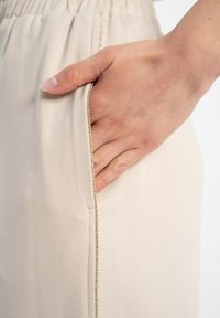 Beżowe spodnie z boczną kieszenią, z złotym akcentem wzdłuż krawędzi. Tkanina wydaje się miękka z gładką teksturą.