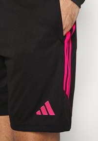 Svarta sportshorts av slätt tyg, med rosa sidostraps och en rosa Adidas-logotyp längst ner till vänster.