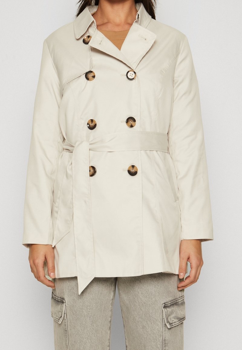 Trench-coat beige en tissu lisse, doté d'un design croisé, de grands boutons et d'une ceinture à nouer à la taille.