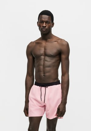 Gespiervde man staand, dragend lichtroze zwemshorts met zwarte tailleband en koordje, tegen een effen witte achtergrond.